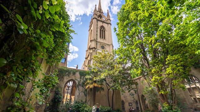 St Dunstan in the East Church Garden with church tower and blue sky in the background, and green trees and benches in the foreground.