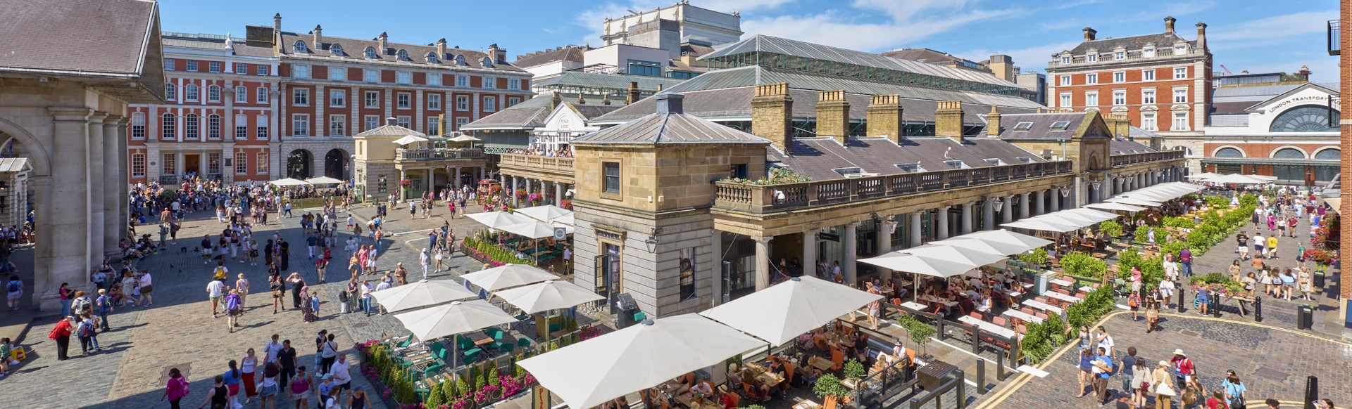 Covent Garden piazza viewed from above with crowds of people walking past building or sitting at outdoor terraces on the square.