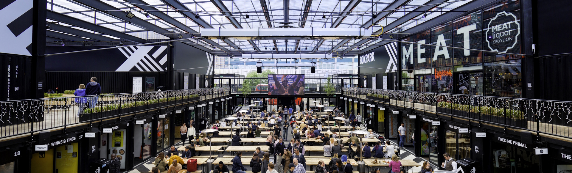 Inside view of Boxpark Croydon with wooden benches and tables, storefronts, glass roof and people throughout the venue.