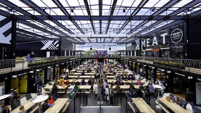Inside view of Boxpark Croydon with wooden benches and tables, storefronts, glass roof and people throughout the venue.