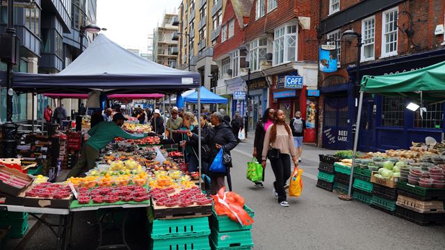 Market food stalls with fruits and vegetables on Surrey Street in Croydon with people walking past.