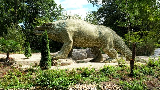 A large green dinosaur statue is standing amongst trees in Crystal Palace Park.