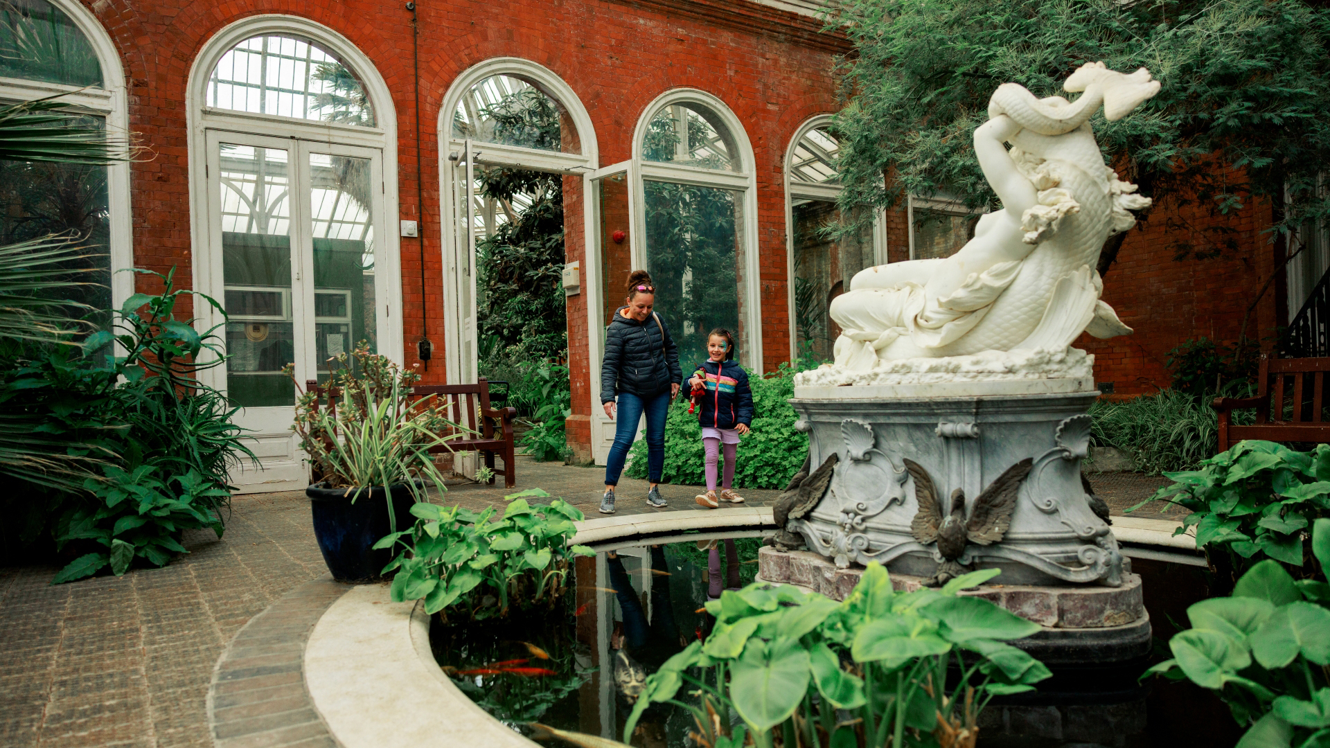A photo of a woman with her young child walking around the winter garden at Eltham Palace with a large water feature in the forgeround