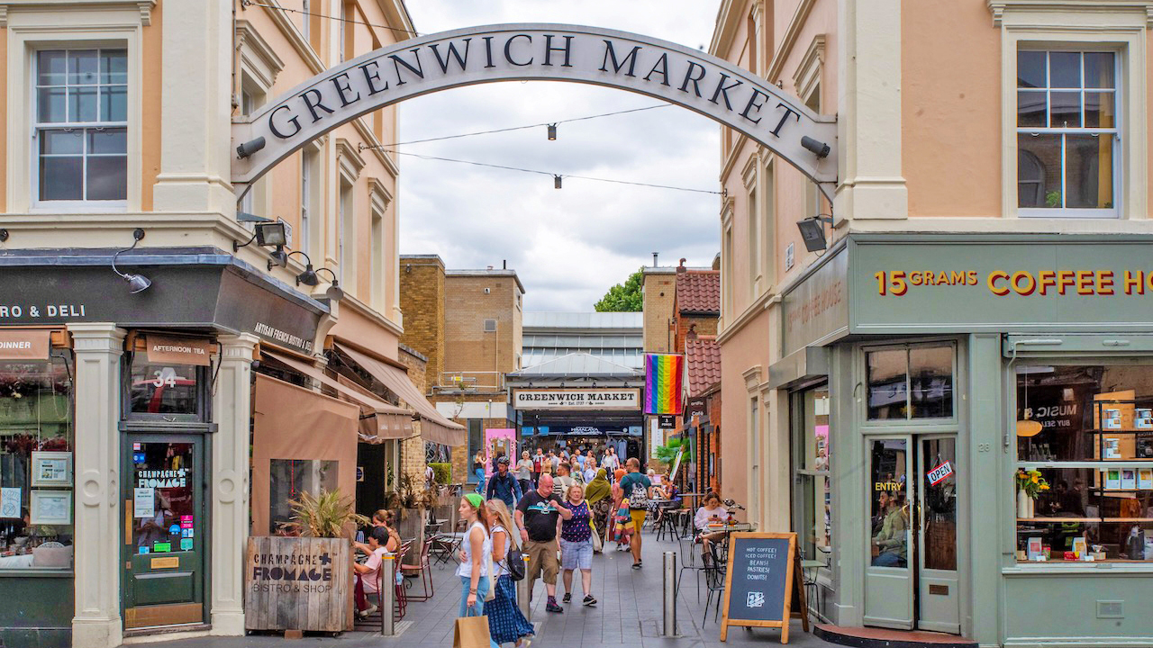A photo of the entrance to Greenwich Market with people walking around holding shopping bags and a sign overhead that reads 