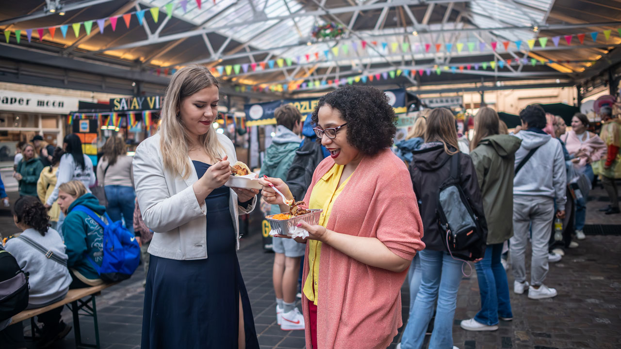 Profitez d'une session shopping a Greenwich Market avant de vous laisser tenter par une sélection aléchante de street food. ©London & Partners/Michael Barrow Deux femmes se trouvent dans la cour du marché de Greenwich et dégustent quelques plats street food.