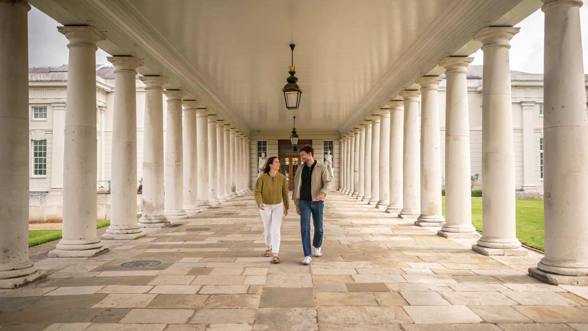 A photo of a man and woman walking in the middle of a walkway in front of the Old Royal Naval College with columns either side of them
