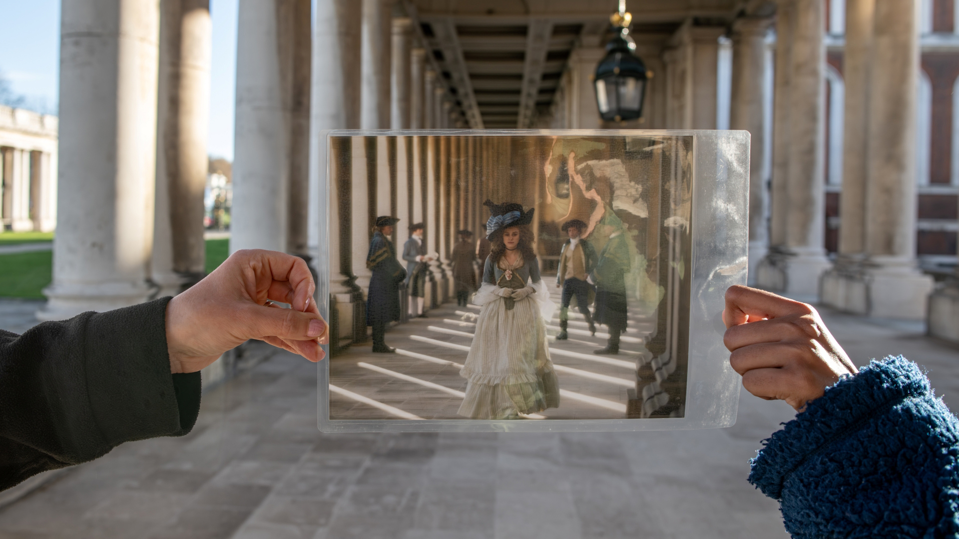 A photo of someone holding up a photo of a still from Bridgerton in front of the site it was filmed at a walkway on the Old Royal Naval College grounds