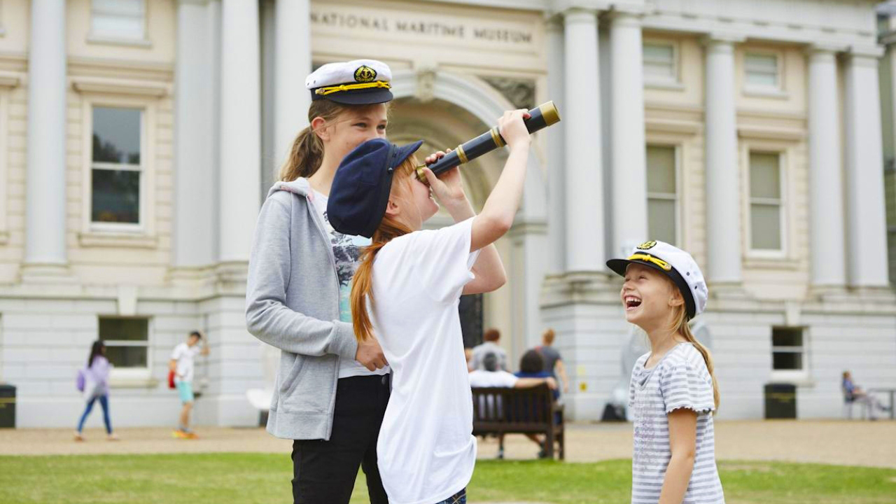 A photo of three children of different ages laughing together outside the National Maritime Museum, each wears a sailor cap and one is looking through a telescope 