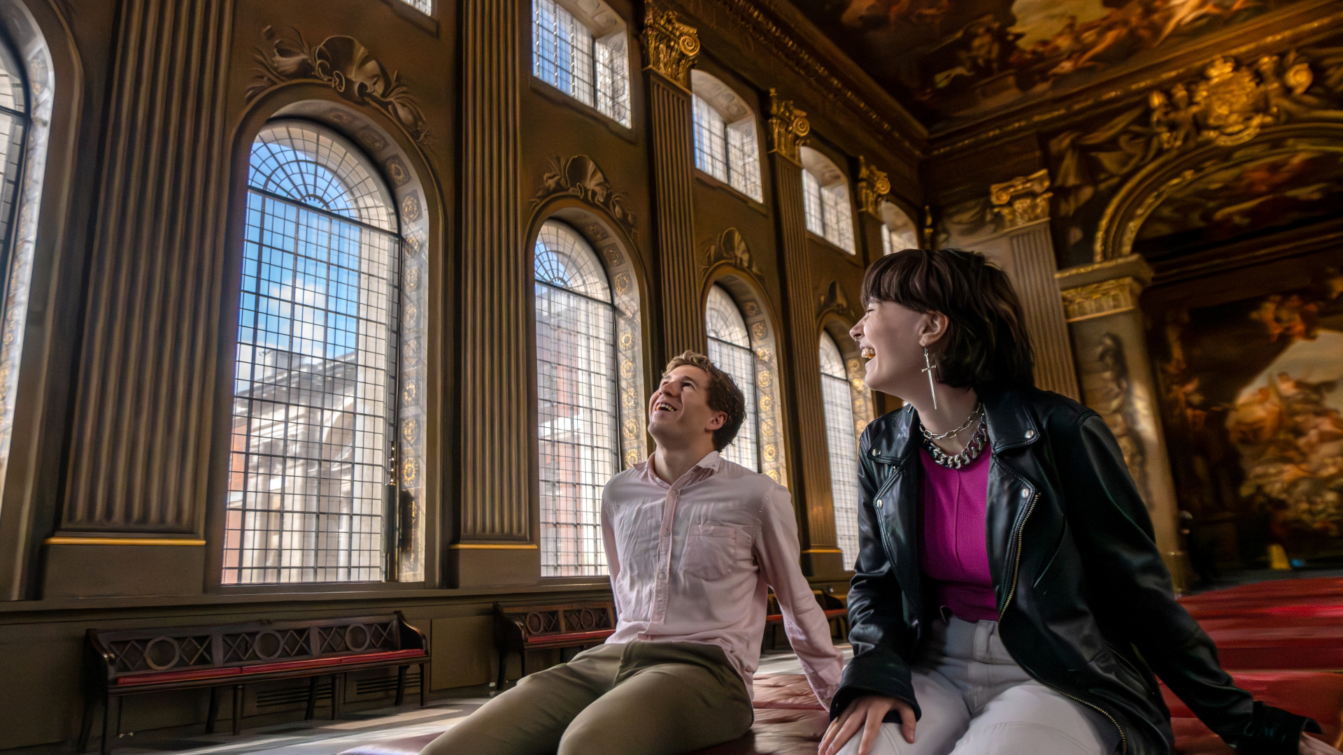 A photo of a man and woman sat down inside the Painted Hall smiling together whilst admiring the painted walls and ceiling