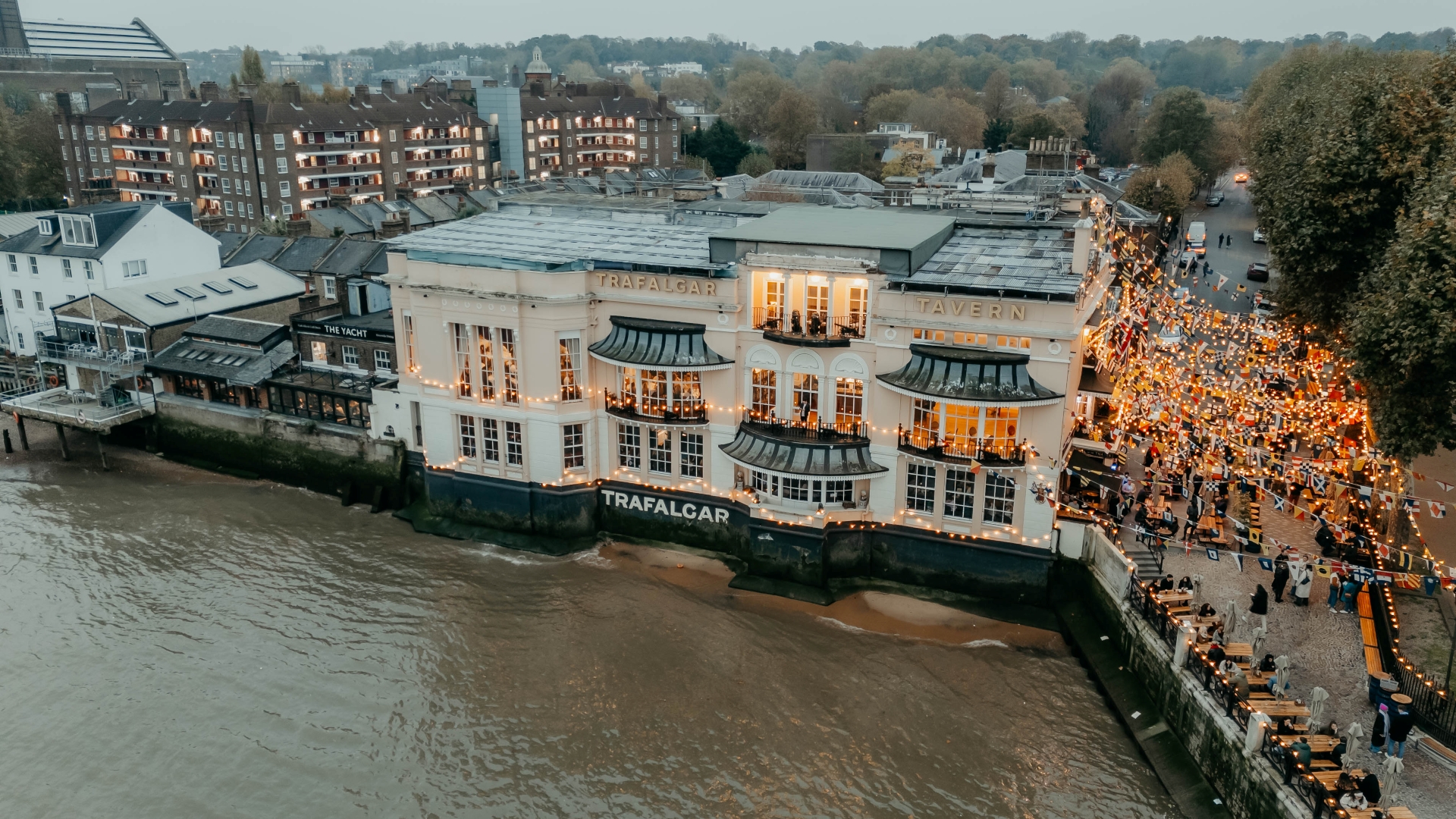 A bird's-eye view of the Trafalgar Tavern riverside pub in Greenwich with warm fairy lights lining the outdoor area and crowds of people stood and sat by the river Thames
