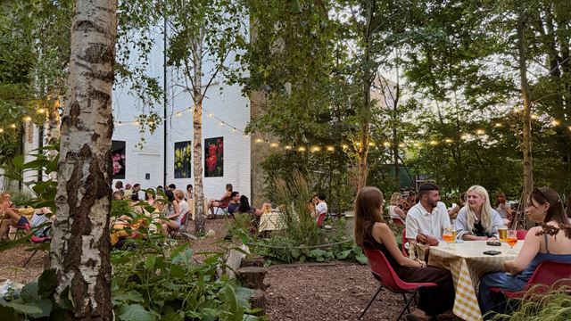 A photo of Dalston Eastern Curve Garden in the evening with groups of people sat on tables and fairy lights hanging overhead