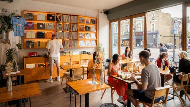 A photo of Next Door Records Two with people sat on wooden tables enjoying small plates and drinks while someone flicks through records at the back of the venue