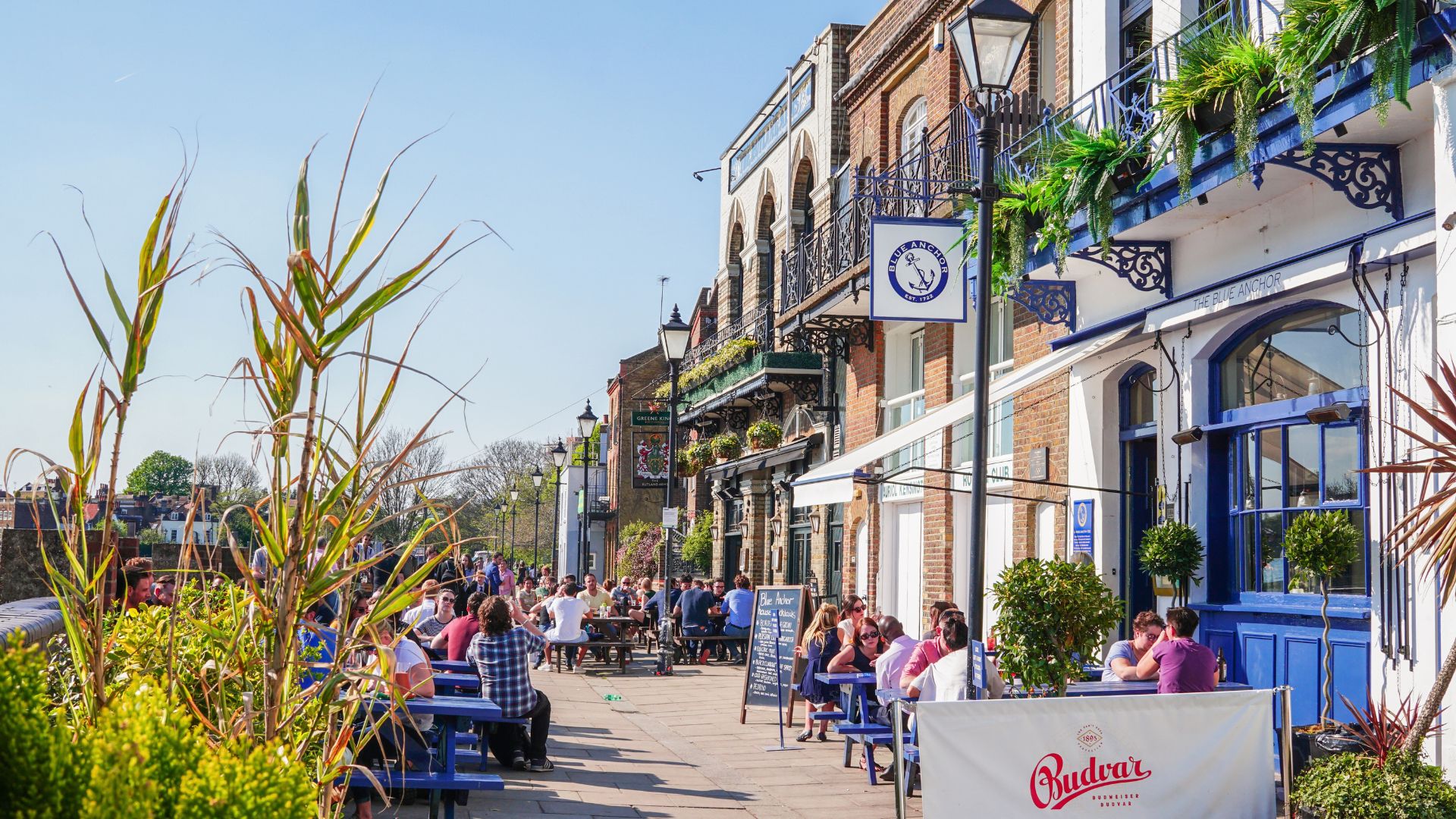 People sitting outside riverside pubs and restaurants in Hammersmith on a sunny day.
