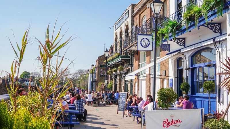 People sitting outside riverside pubs and restaurants in Hammersmith on a sunny day.