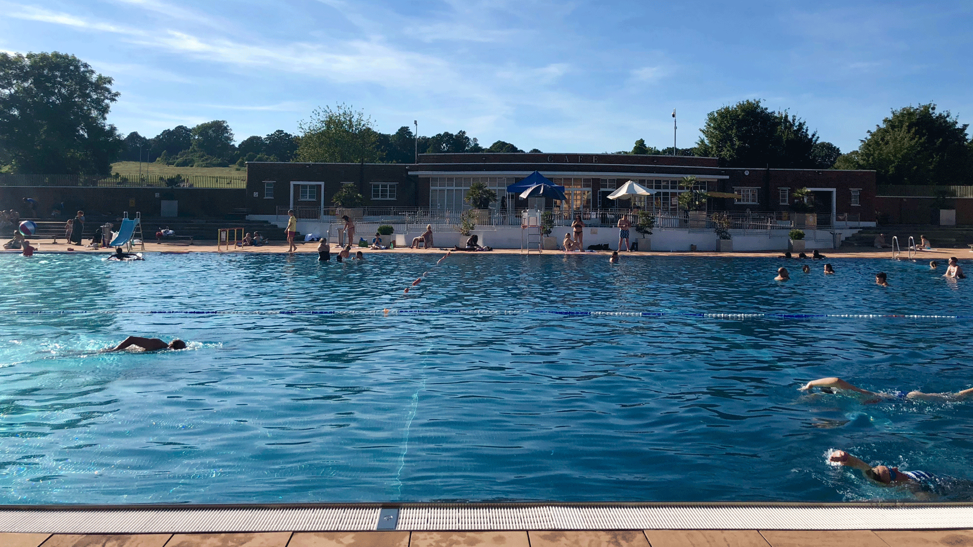 Dive into the refreshing waters at Hampstead Heath Lido. Image courtesy of Shutterstock. People swim in the lido at Hampstead Heath with a view of the park in the background