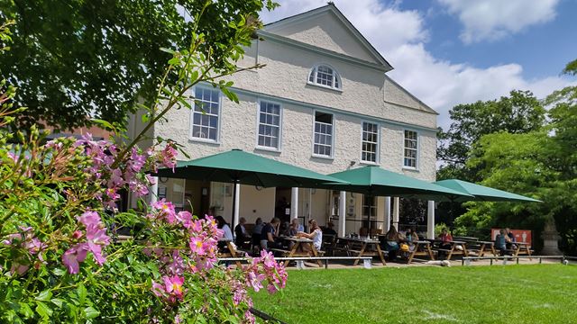 Lauderdale House is shown from the garden side, with trees sporting pink blooms and visitors enjoying refreshments on the terrace of the café.