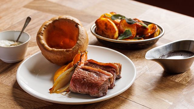 Close up of a beef roast with Yorkshire pudding, vegetables and sauce on the side on a wooden table.