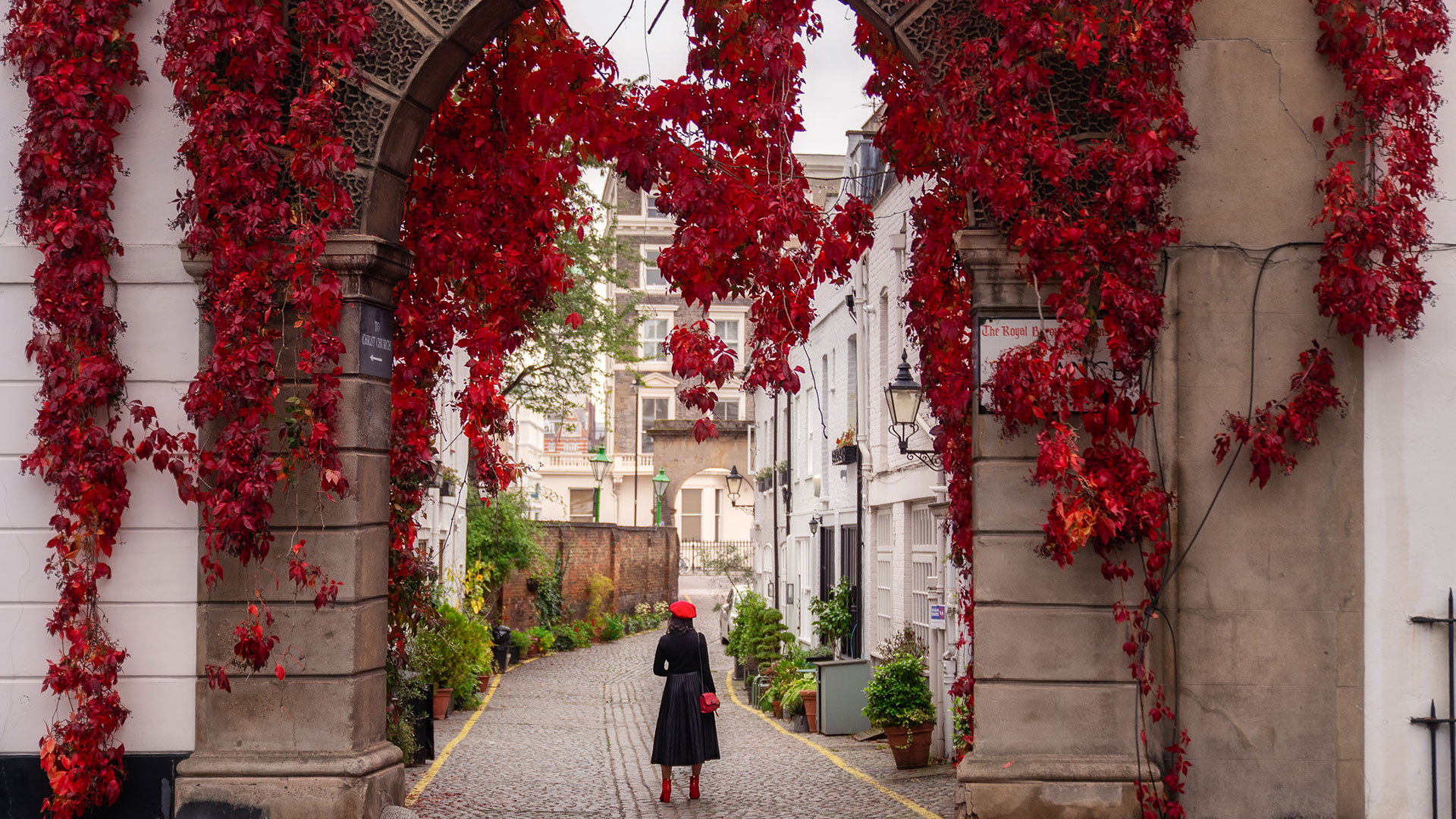A woman dressed in a black coat and a red hat standing in front of Kynance Mews in Kensington, with red flowers hanging from the archway.