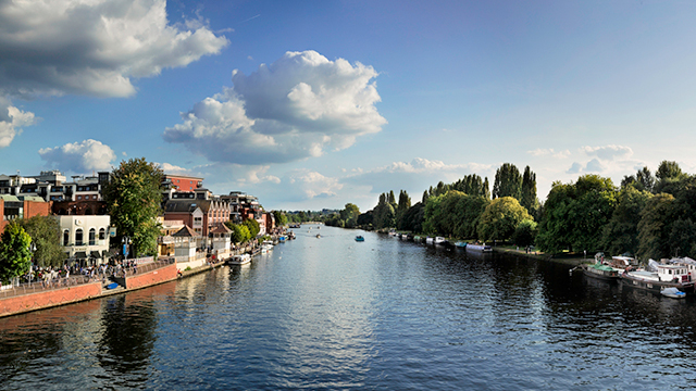 Looking at the river Thames, from a bridge, in Kingston Upon Thames on a sunny day. River boats are moored on the right bank, surrounded by trees; while there are buildings and a walkway on the left bank.