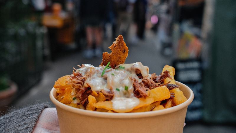A person holding a paper bowl filled with French fries topped with pulled confit duck and melted cheese.