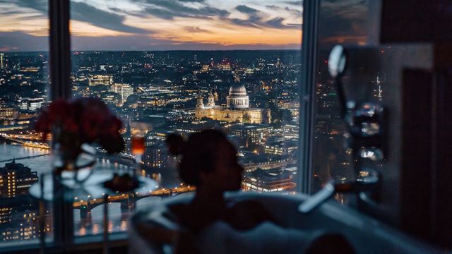 A woman in a bath at night with floor-to-ceiling hotel windows behind her overlooking sweeping London views with St Paul's Cathedral in the distance.