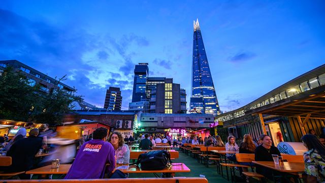 Customers enjoying a drink on wooden benches in Vinegar Yard's outdoor seating area just after sunset, with The Shard and Guy's Hospital in the background.