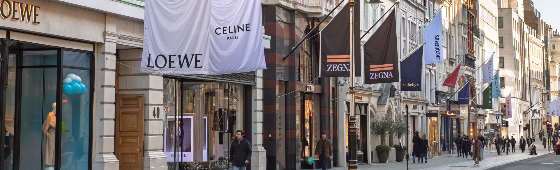 A view along Bon Street with shopfronts and flags showing the branding of designer shops such as Loewe, Celine and Zegna.