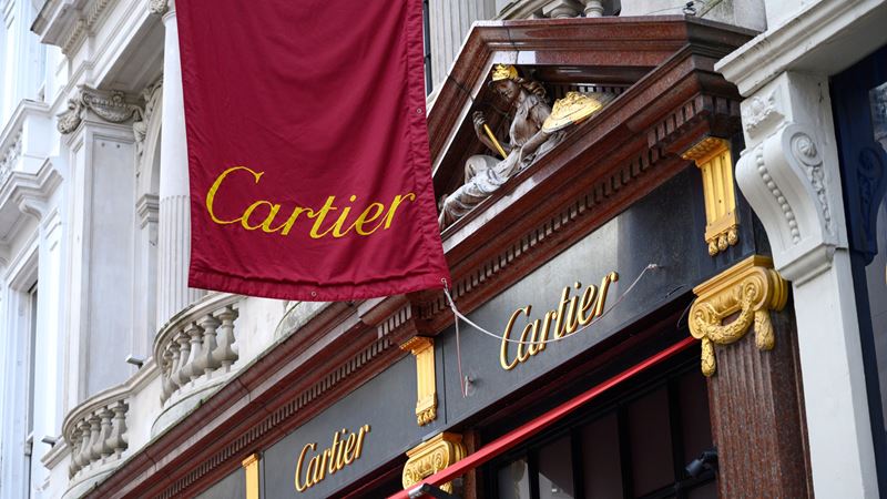 A close-up of Cartier's shopfront on Bond Street, including a red flag with Cartier written in gold, a black hoarding and historic facade.