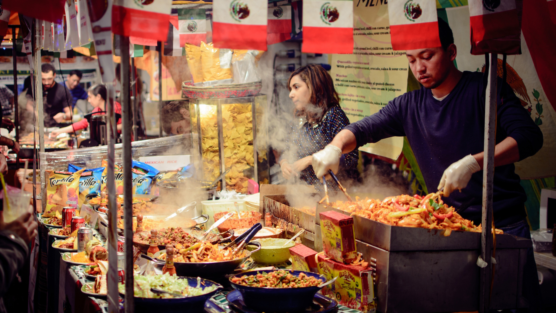 A photo of two cooking and taking orders at a Mexican food stall in Camden Market with lots of vibrant dishes cooking on grills in front of them