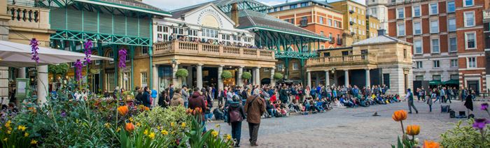 A photo of the Covent Garden piazza with lots of people sat and stood outside watching a performer and plants and flowers in planters in the foreground