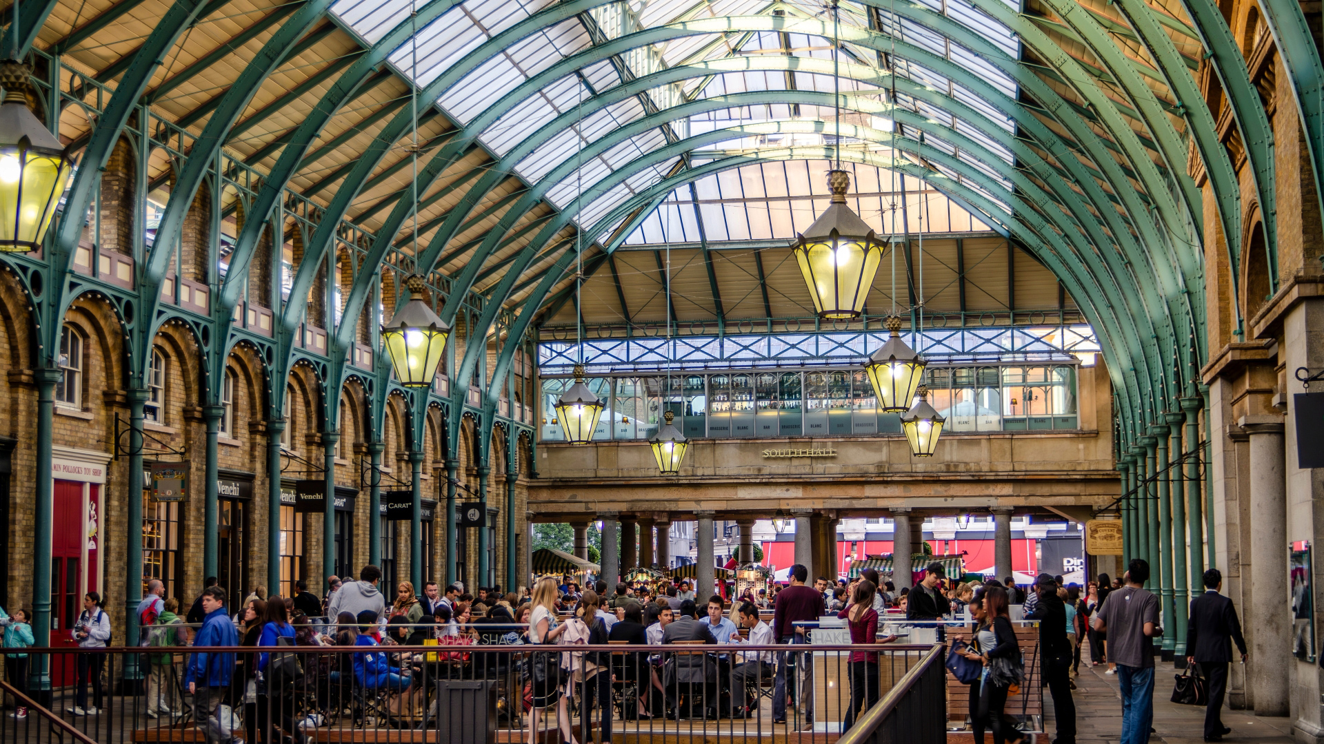 A photo of the Covent Garden piazza with its teal painted beams and lanterns with people walking around in the middle of the building
