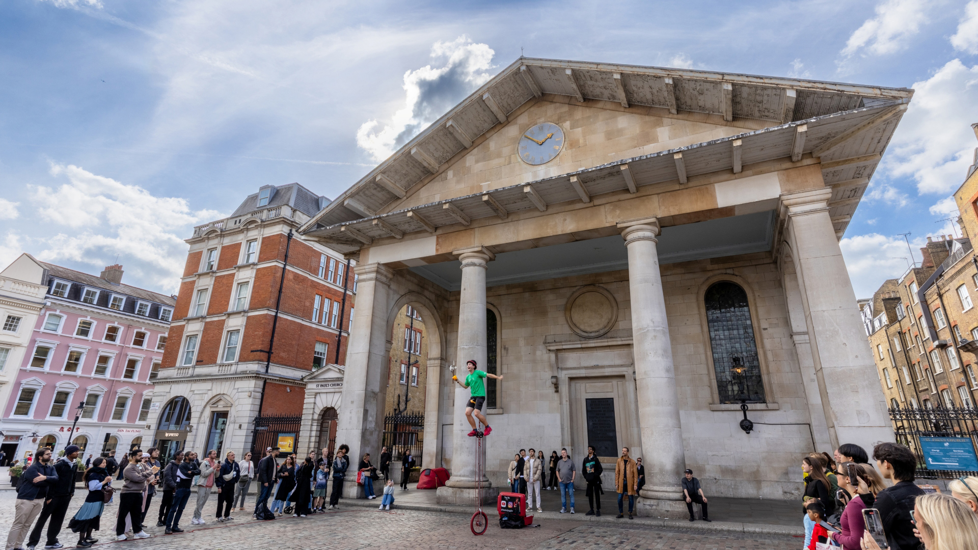 A photo of a man on a giant unicycle juggling in front of crowds of people in the main square of Covent Garden
