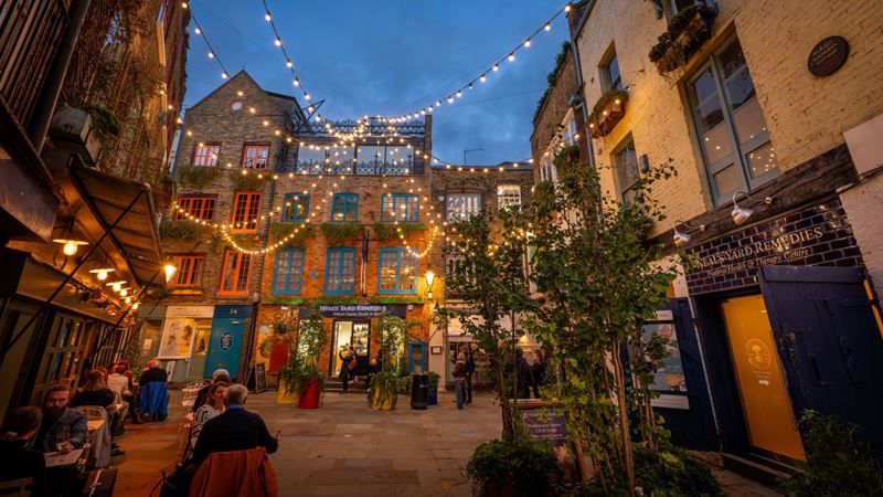 A photo of Neal's Yard at night with colourful shopfronts, people sat outside at tables and fairy lights hanging overhead
