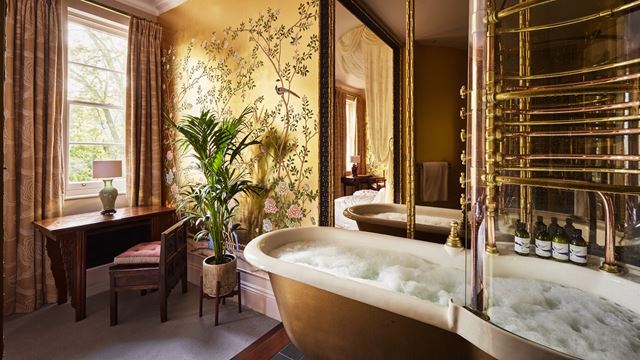 A luxury hotel bathroom with a roll-top bath filled with water and bubbles, yellow wallpaper, a small desk with a chair and a plant by the window.