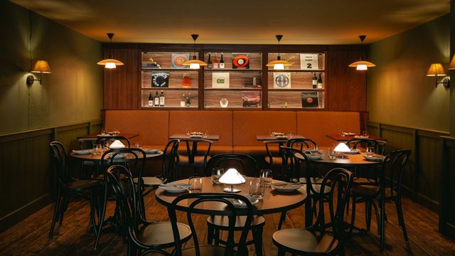Inside darkly lit wine bar and restaurant, with wooden tables and chairs, bottles of wine and vinyls on shelves in the background and hanging lights.