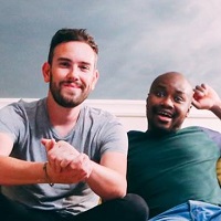 Lloyd and Yaya bloggers of Hand Luggage Only sit on a sofa in front of a grey wall.