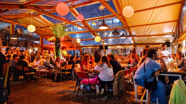People enjoying drinks and conversation inside Peckham Arches with hanging lights and a palm tree in the background.