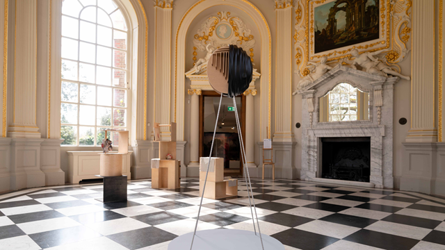 Contemporary artwork displayed in an ornate room at Orleans House Gallery, featuring a black and white chequerboard floor, gilded decorations, a marble fireplace and large windows.