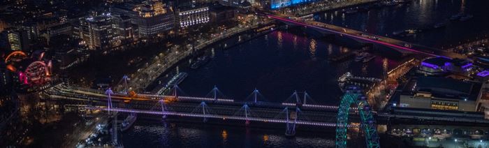 Golden Jubilee and Waterloo Bridges illuminated in pink, purple and blue over the river Thames at night. Around it is either side of the river with buildings and monuments, including the London Eye, all lit up.