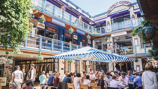 People sitting around communal tables and enjoying food and drinks in the sunshine inside Kingly Court courtyard in Soho.