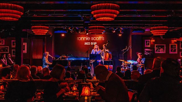 Inside the world-famous Ronnie Scott's jazz bar in London with people listening to live musicians in a darkly lit room filled with red lights, lamps and banquettes.