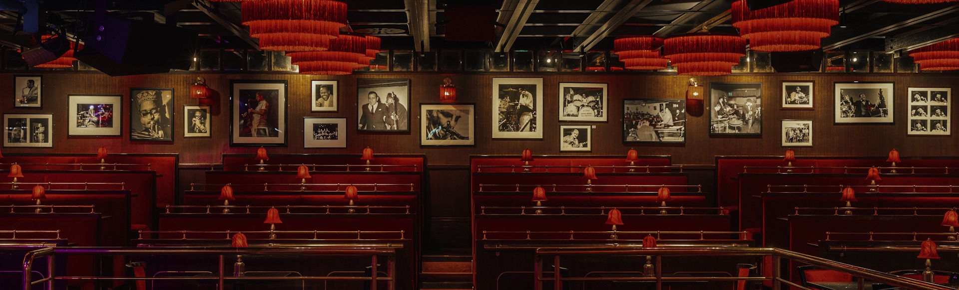 The red-lit interior of Ronnie Scott's jazz club, with black and white pictures of musicians on the wall and rows of bench seating.