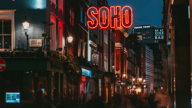 A Soho street at night with a red neon sign saying Soho at the top of the picture.