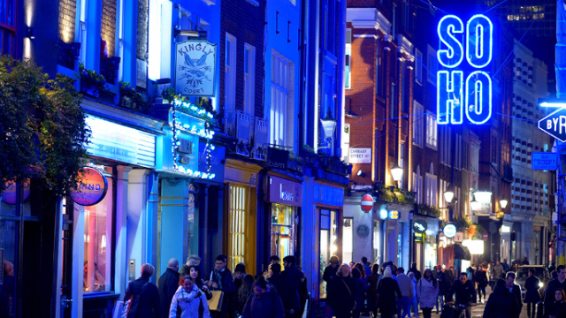 Soho street with people at night with a blue neon sign on a building that says Soho.