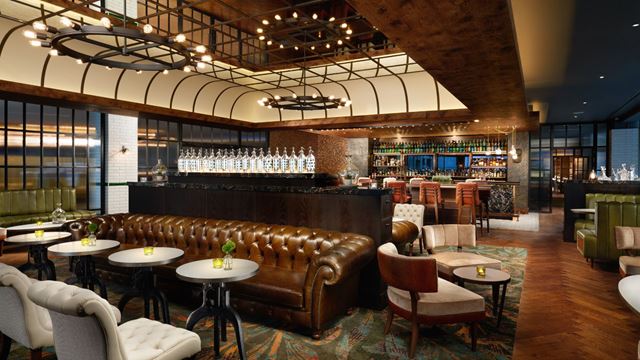 an art deco themed hotel lounge with plush brown leather furniture, spotlight lighting, green booths and a bar in the background