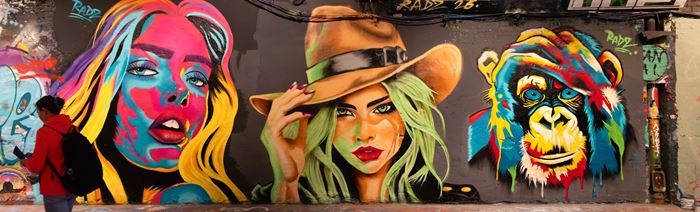 A woman walking past a wall decorated with brightly coloured street art featuring the faces of two women and a chimpanzee.
