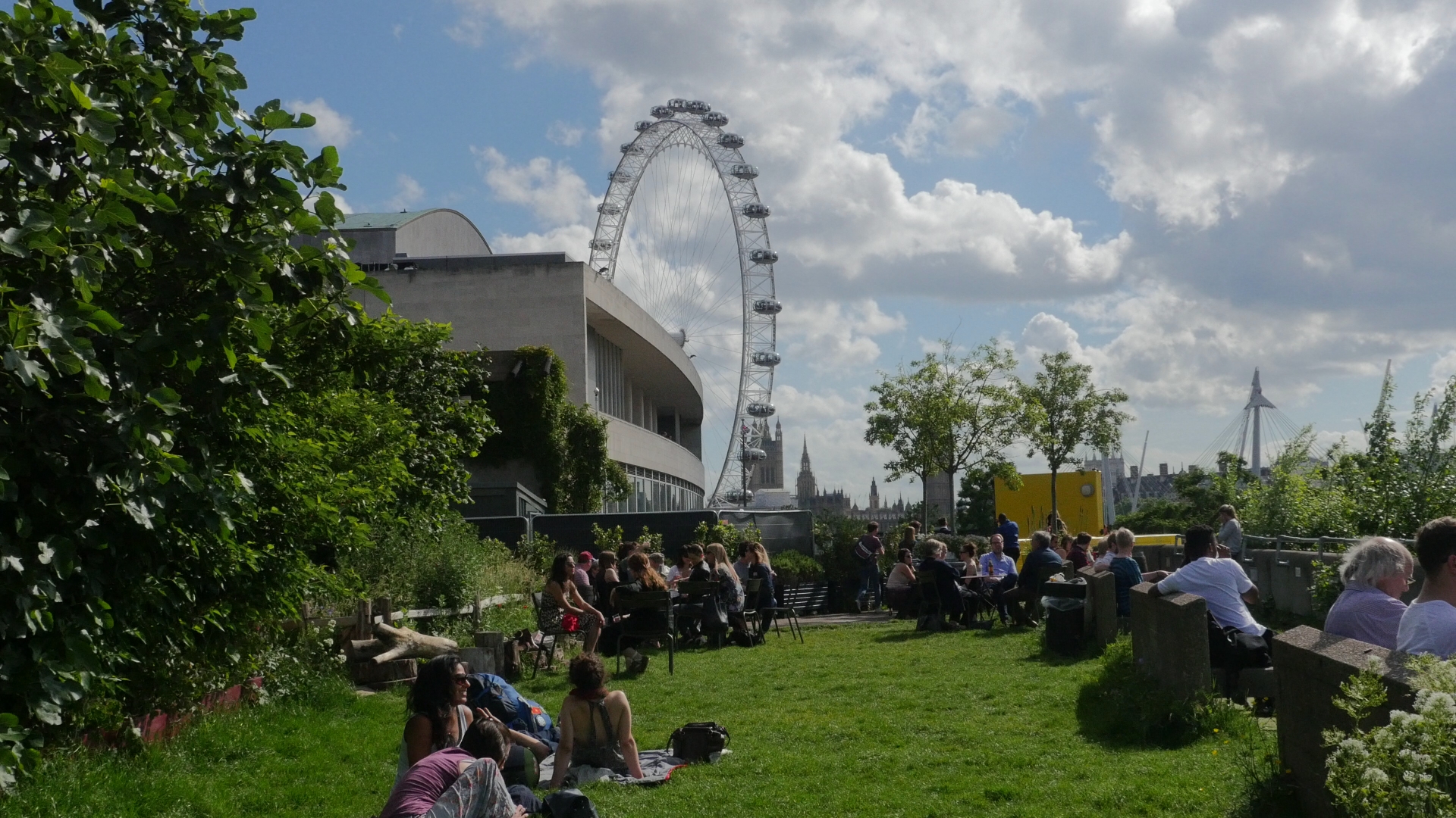 People relaxing on the grass on a bright day at Queen Elizabeth Hall Roof Garden, with the London Eye in the background.