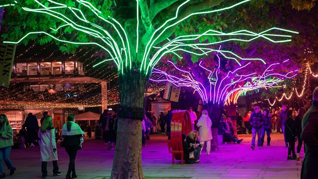 A stretch of the South Bank with Christmas lights, market stalls, and trees lit up with green and purple LEDs.