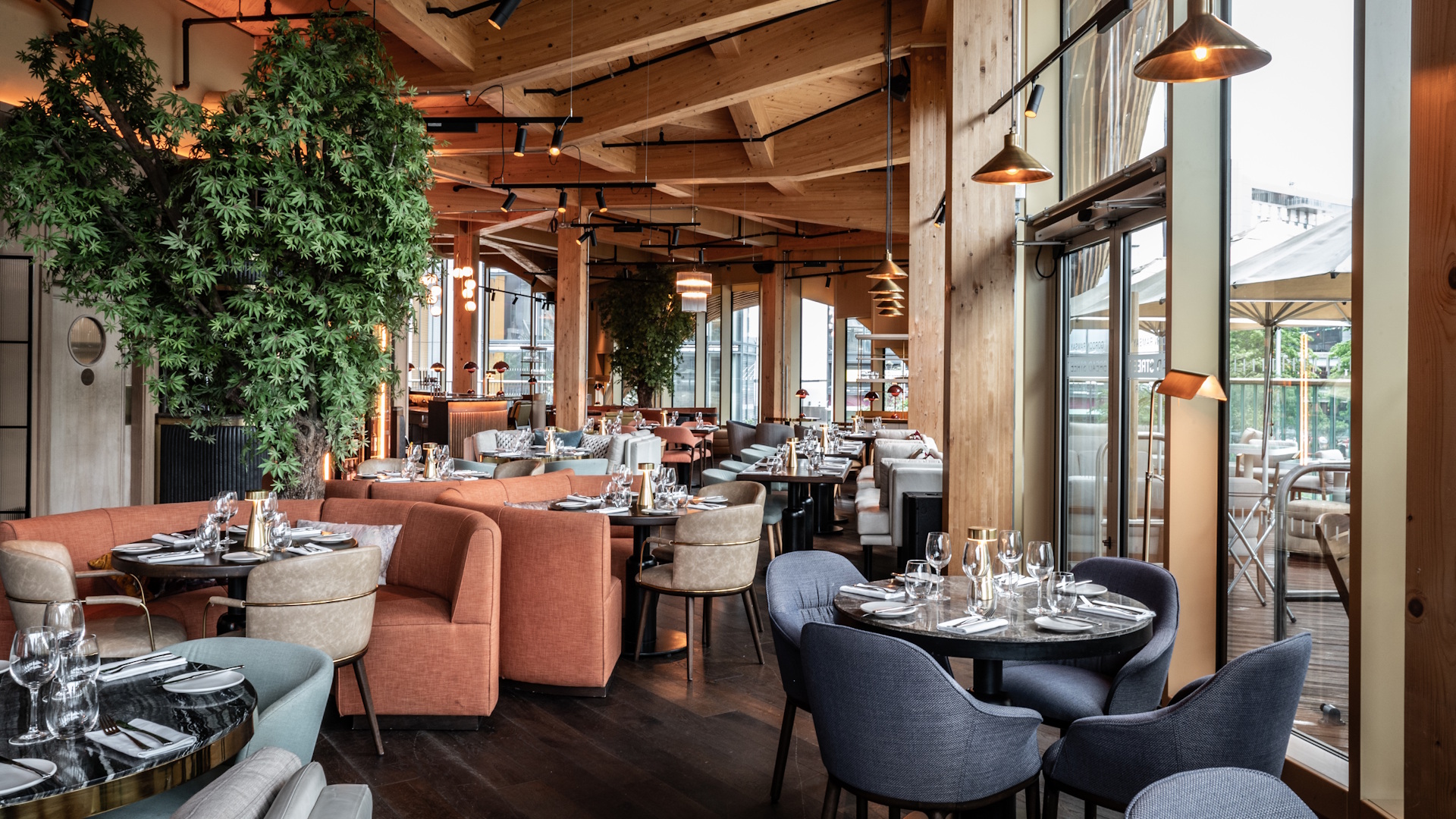 Interior dining room at Bread Street Kitchen in Stratford, London, featuring floor-to-ceiling windows, orange and grey seating, industrial-style lighting, wooden ceiling and hanging plants.