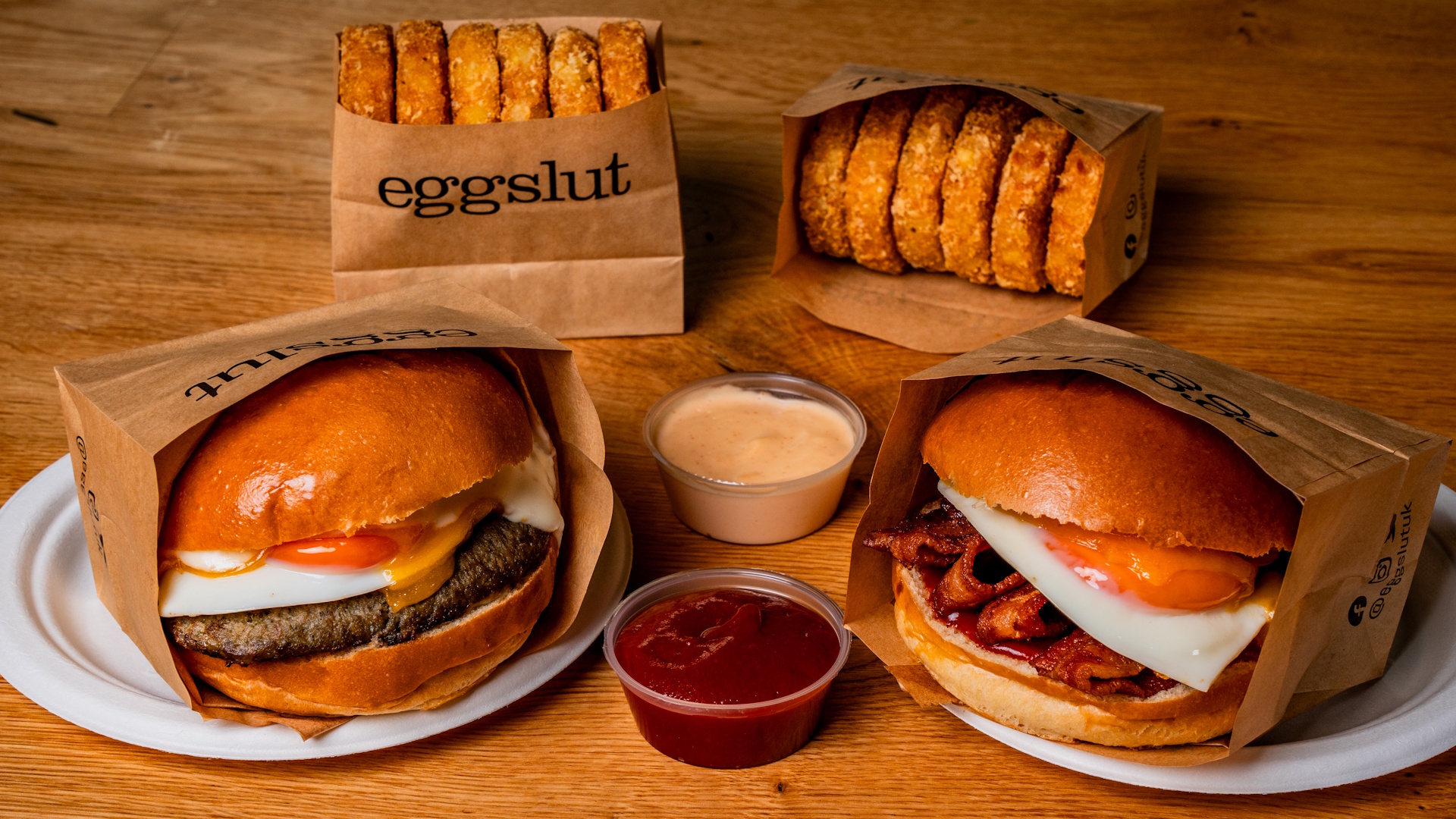 Two brioche buns from eggslut with a bright orange fried egg on top of bacon and a beef patty, with sauces and sides on the wooden table.